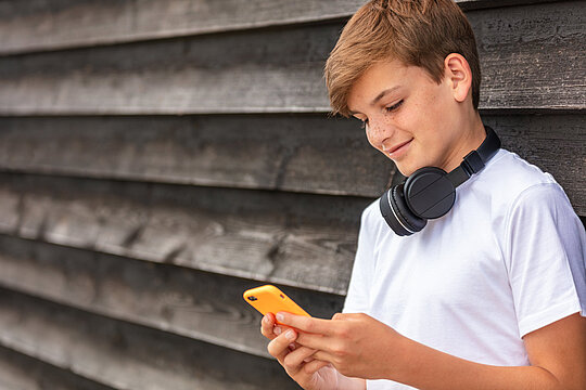 Ein kleiner Junge trägt Kopfhörer und hält ein Mobiltelefon an eine Holzwand.